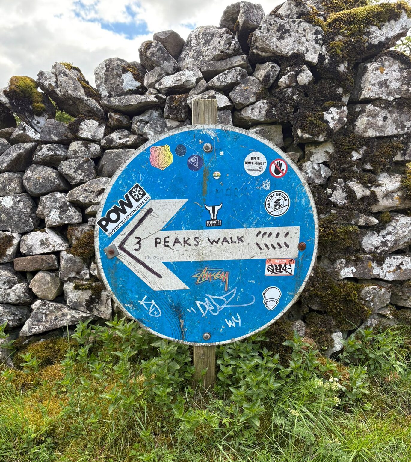 A blue circular wayfinding sign stands in front of a dry stone wall in a rural setting. The sign features an arrow and the handwritten words “3 Peaks Walk”, along with numerous stickers and markings. Grass and wild plants grow around the base of the sign, while moss-covered stones form the wall behind it.