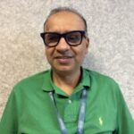 Paresh, North London SLC member leans against a light grey wall at the annual SLC Conference. He wears dark rimmed glasses, a vibrant green polo shirt, and has a Sight Loss Councils lanyard around his neck.