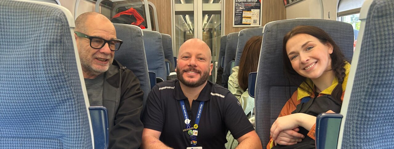 Mick, Sight Loss Council member, seated inside a train during a Try a Train event, alongside a Northern staff member and Community Rail Lancashire volunteer. All are smiling towards the camera.