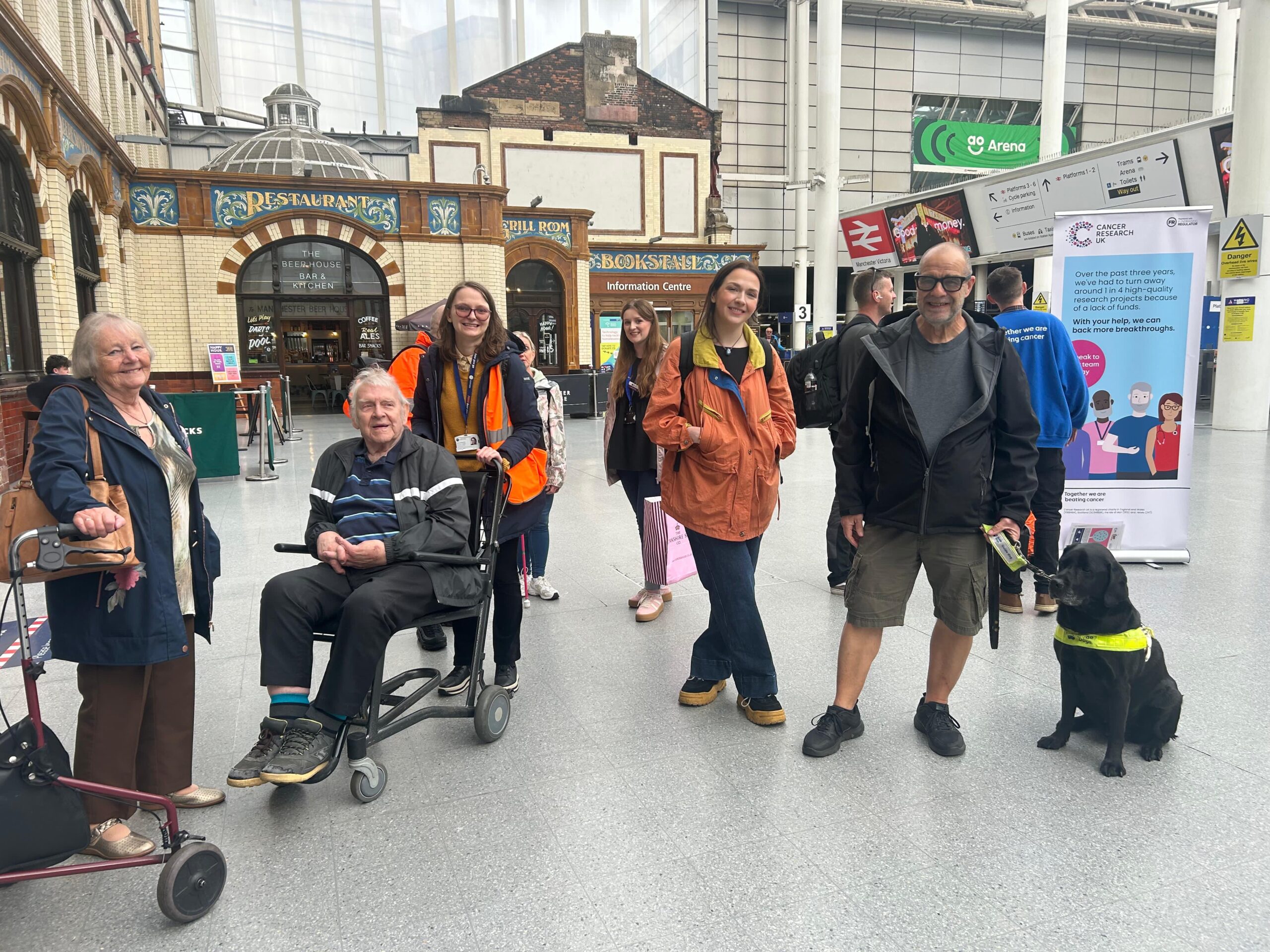 A group of blind and partially sighted participants and volunteers at Manchester Victoria station during a Try a Train event, including a guide dog, with station facilities visible in the background.