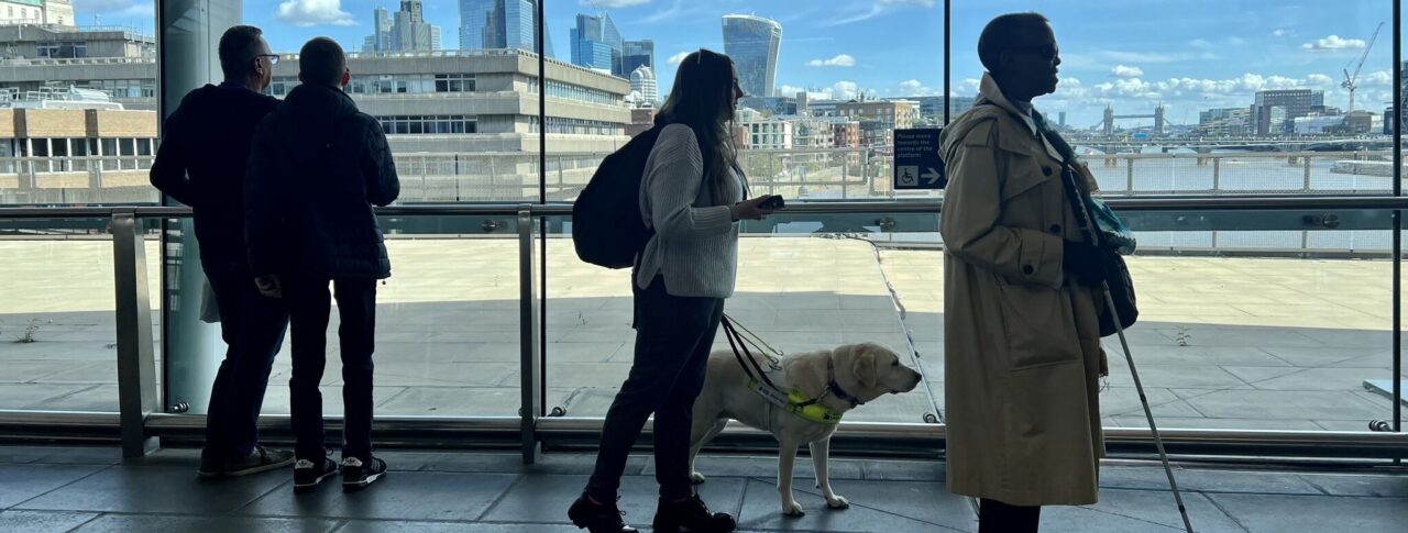 SLC staff and volunteers taking part in an accessibility assessment at London Blackfriars station stand near large windows overlooking the River Thames. One participant is with a guide dog and another uses a long cane as the group explores the station environment as part of the Your Station, Your Community project.