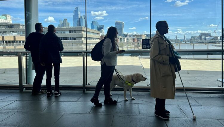 SLC staff and volunteers taking part in an accessibility assessment at London Blackfriars station stand near large windows overlooking the River Thames. One participant is with a guide dog and another uses a long cane as the group explores the station environment as part of the Your Station, Your Community project.