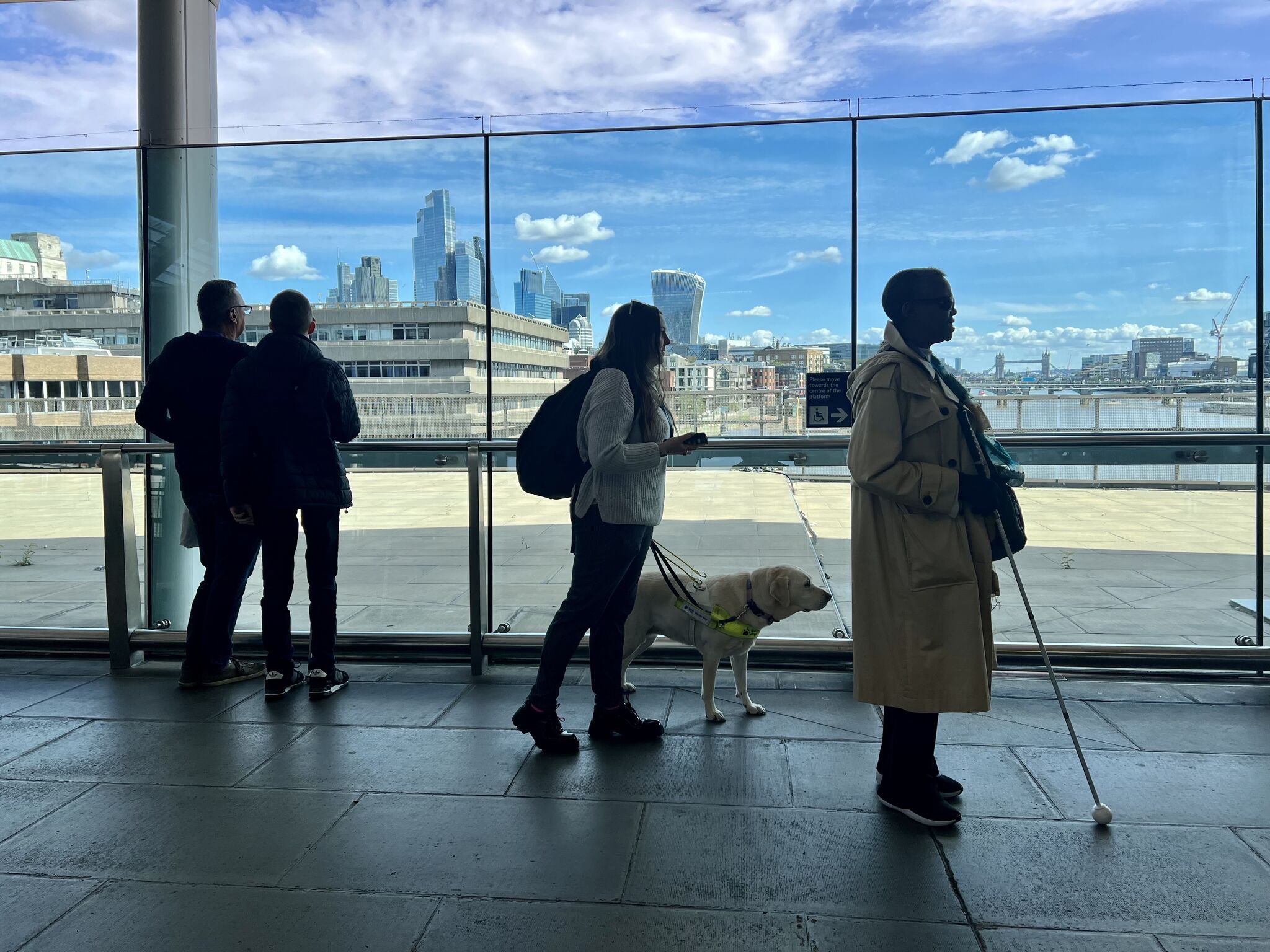 SLC staff and volunteers walk along a platform at London Blackfriars station, in front of large windows overlooking the River Thames. One participant is with a guide dog and another uses a long cane as the group explores the station environment as part of the Your Station, Your Community project.