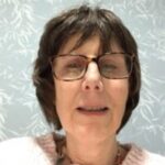 Claire, a Gloucestershire and Swindon Sight Loss Council member, pictured in a head-and-shoulders portrait. Claire is wearing glasses and a light-coloured knitted jumper, seated indoors in front of a pale patterned wall.