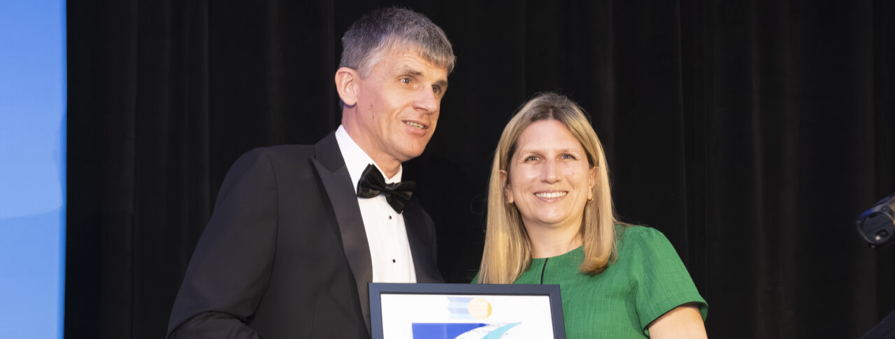 Dave Smith, Engagement Manager for South East England, stands on stage with a CIHT Awards presenter. Dave is wearing a black tuxedo and holding a long cane, while the presenter is wearing a green dress. A dark curtain forms the backdrop, with stage lighting visible to one side. Dave and the presenter are holding a framed award and trophy for the camera.