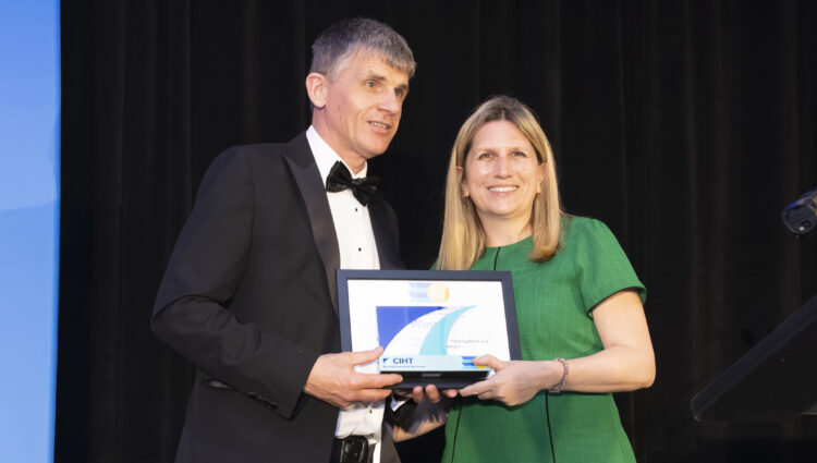 Dave Smith, Engagement Manager for South East England, stands on stage with a CIHT Awards presenter. Dave is wearing a black tuxedo and holding a long cane, while the presenter is wearing a green dress. A dark curtain forms the backdrop, with stage lighting visible to one side. Dave and the presenter are holding a framed award and trophy for the camera.