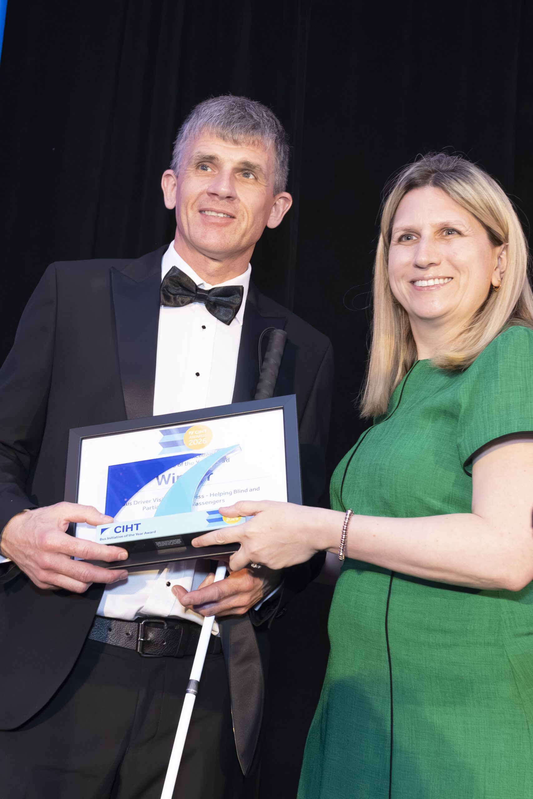 Dave Smith, Engagement Manager for South East England, stands on stage with a CIHT Awards presenter. Dave is wearing a black tuxedo and holding a long cane, while the presenter is wearing a green dress. A dark curtain forms the backdrop, with stage lighting visible to one side. Dave and the presenter are holding a framed award and trophy for the camera.