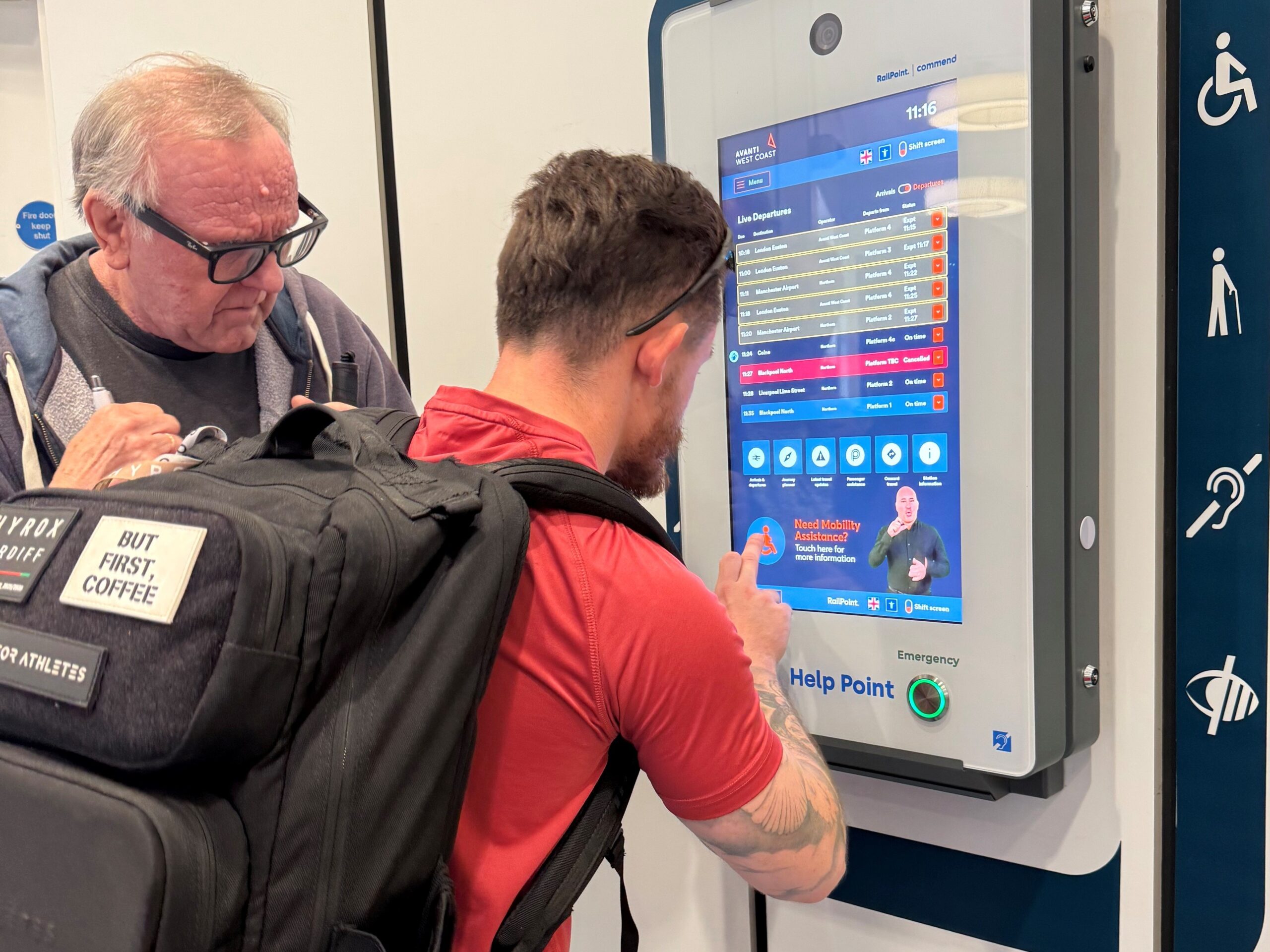 David Parfett, Sight Loss Council Coordinator for North West England, and Sight Loss Council member Michael review a passenger assistance help point during a rail accessibility assessment. The help point displays live train information, accessibility features and mobility assistance options. Accessibility symbols for wheelchair users, hearing assistance and visual impairment are visible on the unit.