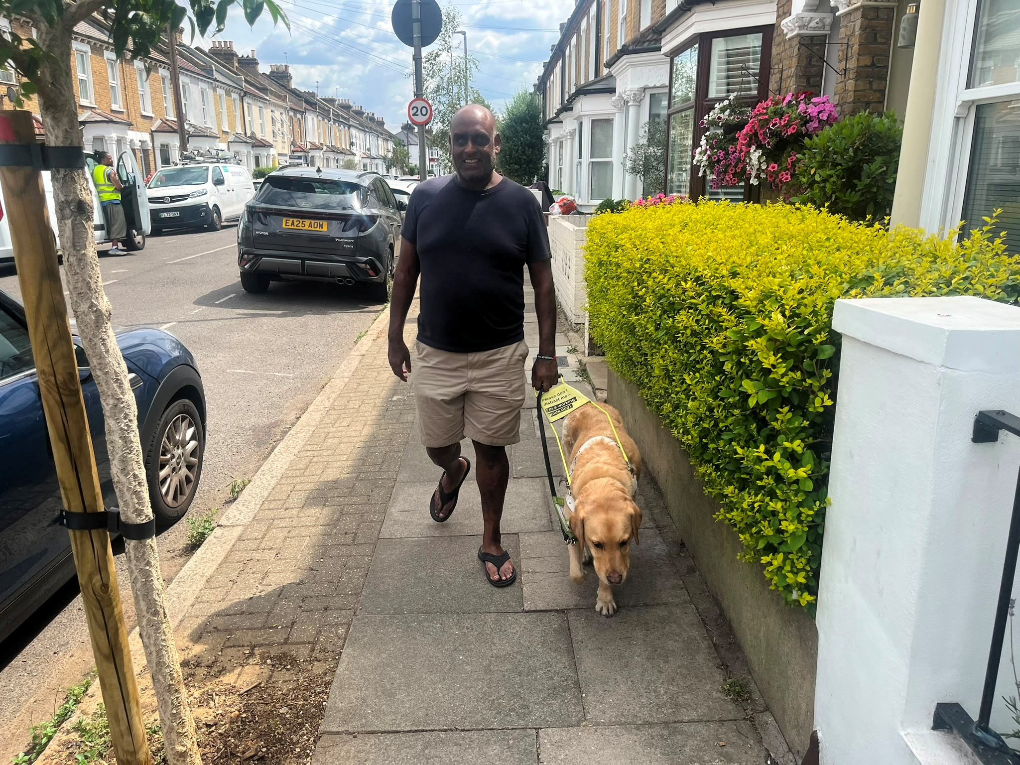 South West London Sight Loss Council member Haren walks along a residential pavement with guide dog Addie. Addie is wearing a guide dog harness and leads the way past parked cars and terraced houses.