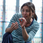Holly, a West of England Sight Loss Council member, sits indoors in front of tall stained-glass windows. Holly is leaning forward and gesturing with both hands while speaking during a discussion or activity.
