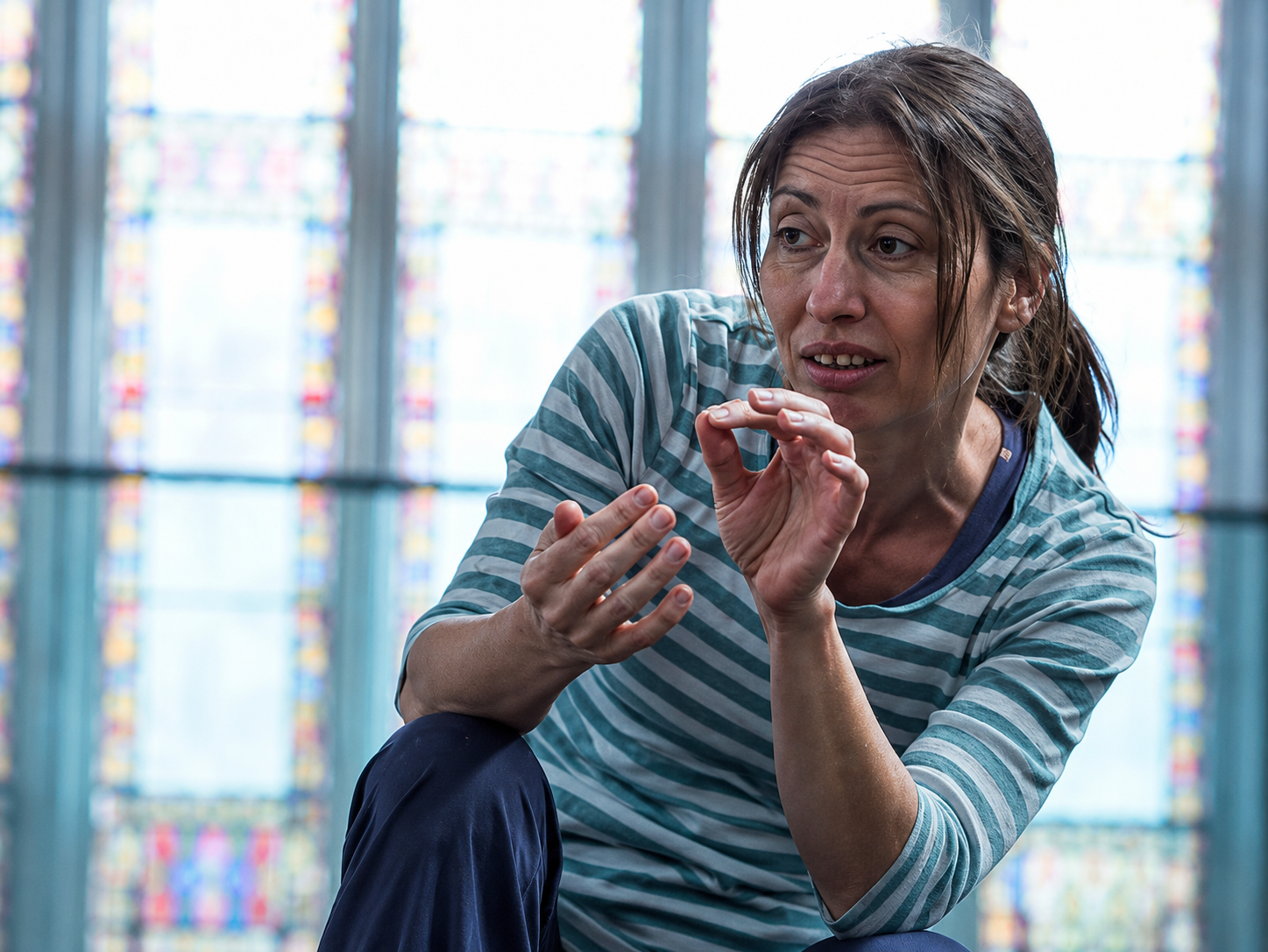 Holly, a West of England Sight Loss Council member, sits indoors in front of tall stained-glass windows. Holly is leaning forward and gesturing with both hands while speaking during a discussion or activity.