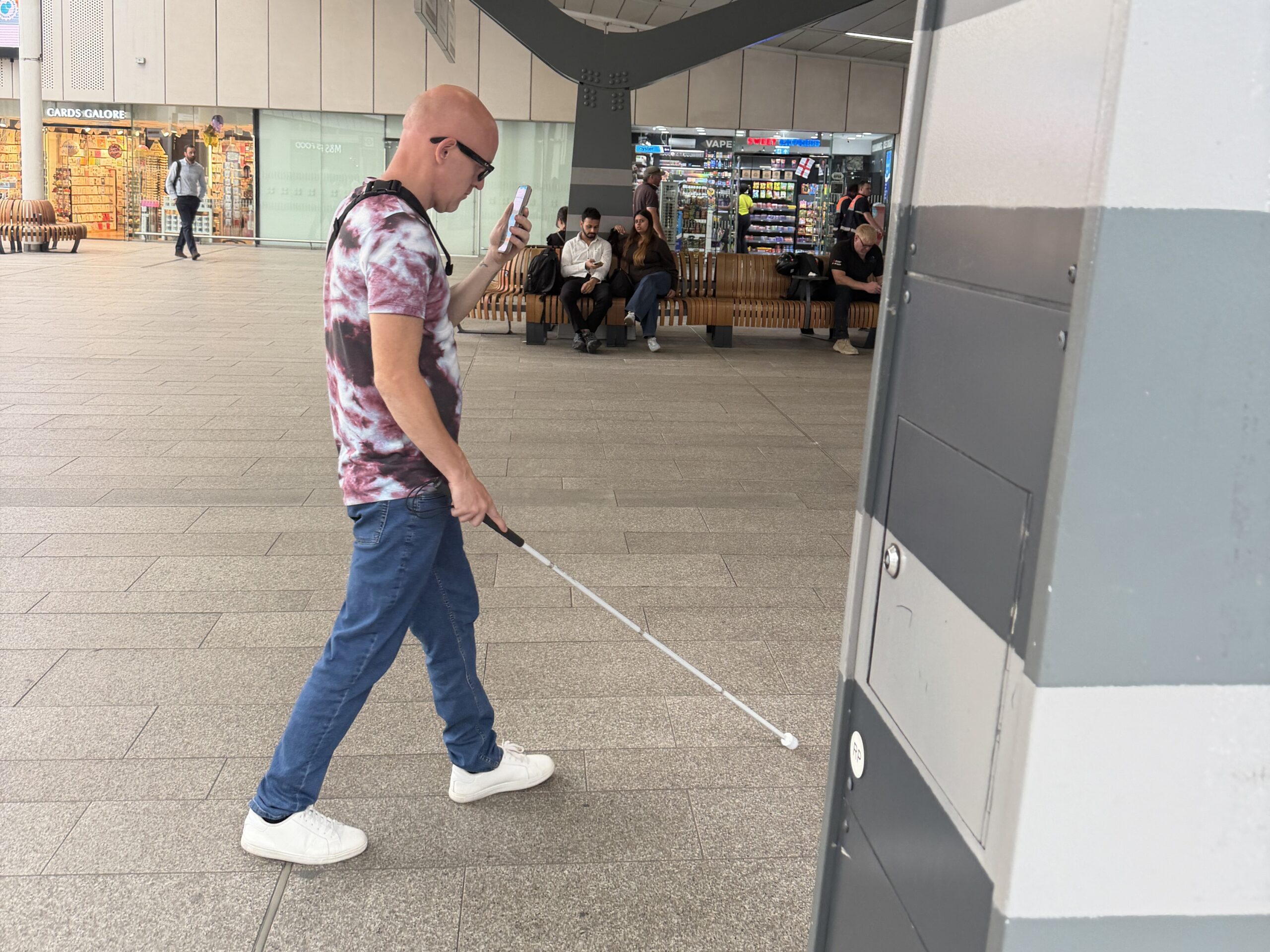 Josh, a Sussex SL C member, navigates a station concourse whilst using a navigation app. He walks with his long cane in one hand, and his smart phone raised in the other.
