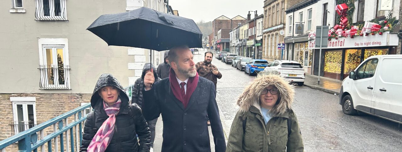 North West England Engagement Manager Kelly Barton, Greater Manchester Sight Loss Council volunteer Hayley Kellard and local MP Jonathan Reynolds take part in an accessibility walkaround in Stalybridge, highlighting barriers faced by blind and partially sighted people. The group walks through the town centre in heavy rain. Jonathan holds a black umbrella up as they walk.