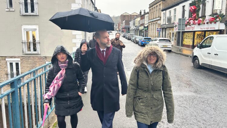 North West England Engagement Manager Kelly Barton, Greater Manchester Sight Loss Council volunteer Hayley Kellard and local MP Jonathan Reynolds take part in an accessibility walkaround in Stalybridge, highlighting barriers faced by blind and partially sighted people. The group walks through the town centre in heavy rain. Jonathan holds a black umbrella up as they walk.