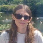 Lauren Eade headshot. Lauren has long brown hair and is wearing sunglasses and a white t-shirt sitting in a garden in the sun.