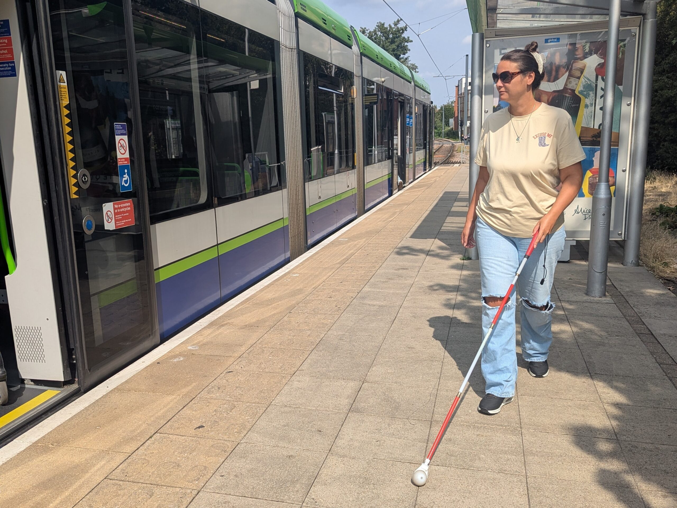 Nikki, South West London SLC member, walks alongside a tram at Wimbledon Station with her long cane. A shelter is shown in the background.