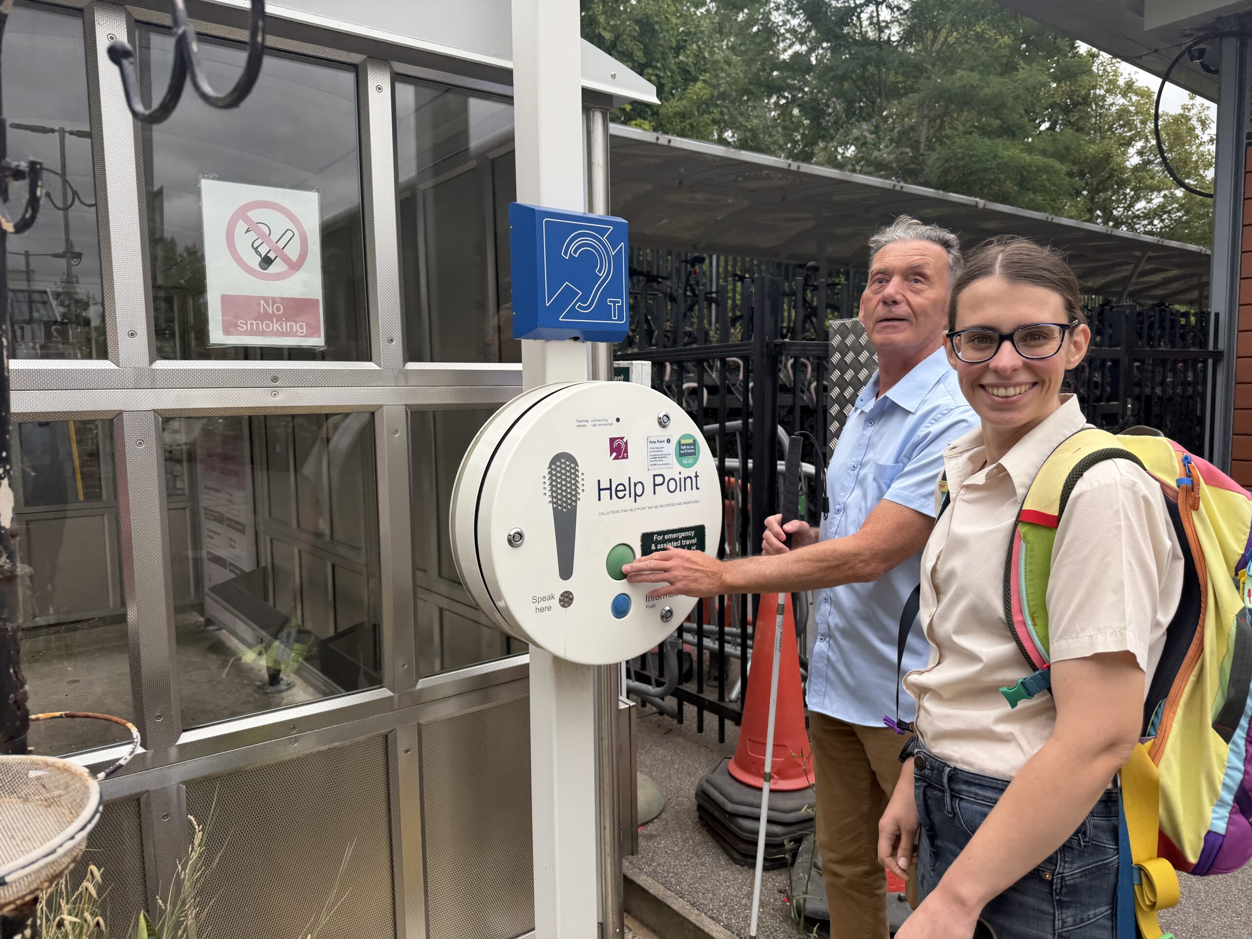 Paul, Sussex SLC member, and Lauren, SLC Coordinator for South East England, stand beside a help point on a station platform. Paul's hand reaches out and presses a large green button, he holds his long cane in his other hand. Both are smiling at the camera.