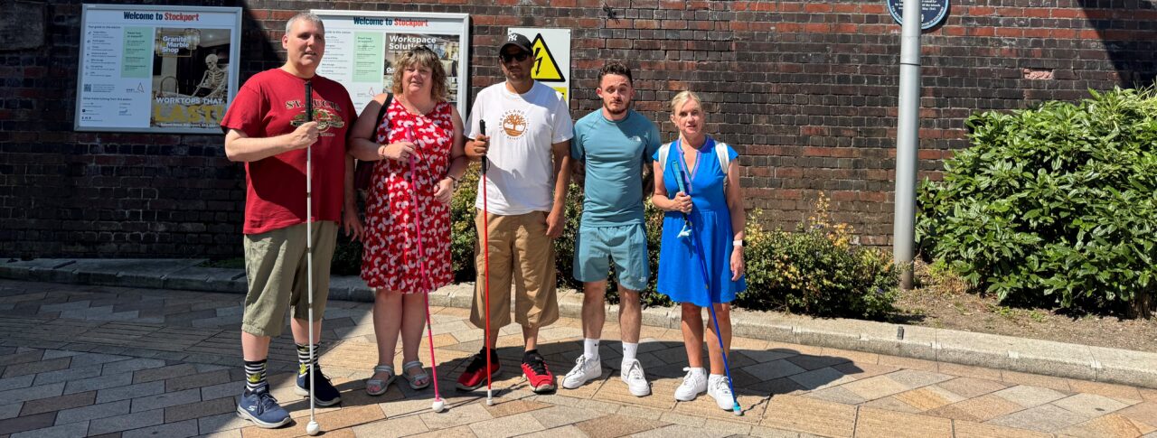 Greater Manchester Sight Loss Council staff and volunteers stand on outside Stockport railway station beneath a large station sign. Four members hold long canes, including white, pink and blue canes, while posing in bright sunshine. The group stands in front of a brick station wall with station information boards and landscaping visible nearby.
