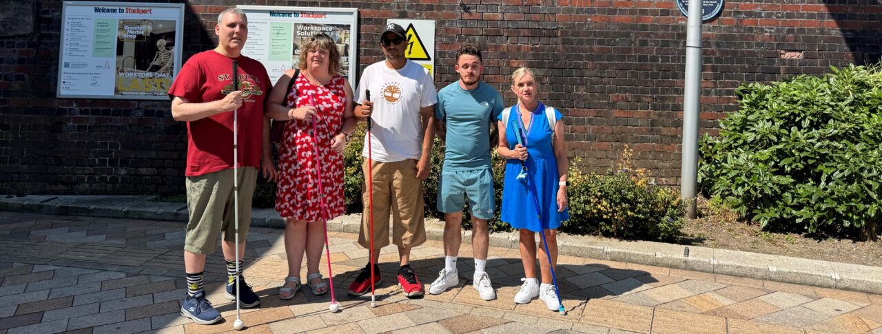 Greater Manchester Sight Loss Council staff and volunteers stand on outside Stockport railway station beneath a large station sign. Four members hold long canes, including white, pink and blue canes, while posing in bright sunshine. The group stands in front of a brick station wall with station information boards and landscaping visible nearby.