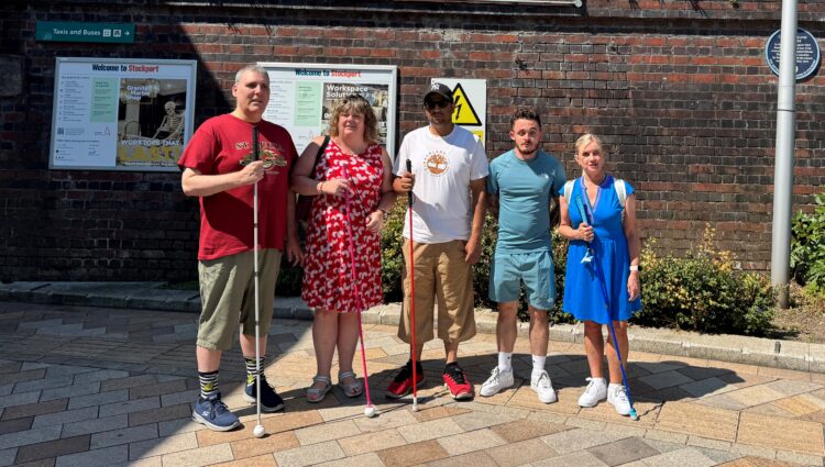 Greater Manchester Sight Loss Council staff and volunteers stand on outside Stockport railway station beneath a large station sign. Four members hold long canes, including white, pink and blue canes, while posing in bright sunshine. The group stands in front of a brick station wall with station information boards and landscaping visible nearby.