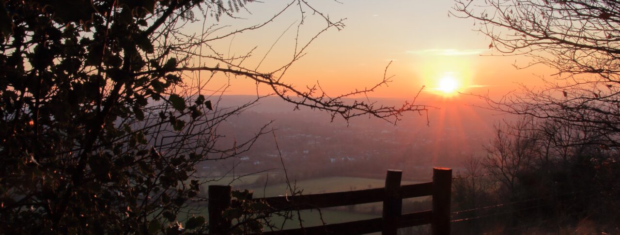 A scenic sunset view from Box Hill in Surrey. The sun is low on the horizon, casting a warm orange glow across the sky and landscape below. Tree branches frame the photo on both sides, and a wooden fence stands in the foreground, overlooking the valley. In the distance, hills and fields appear under the fading daylight.