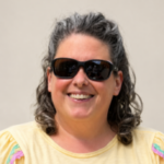A head shot of Beth, a Tyne and Wear SLC member. She is stood against a cream background and has dark, shoulder-length, wavy hair. She is wearing dark sunglasses and a pale yellow t-shirt. She is smiling at the camera.