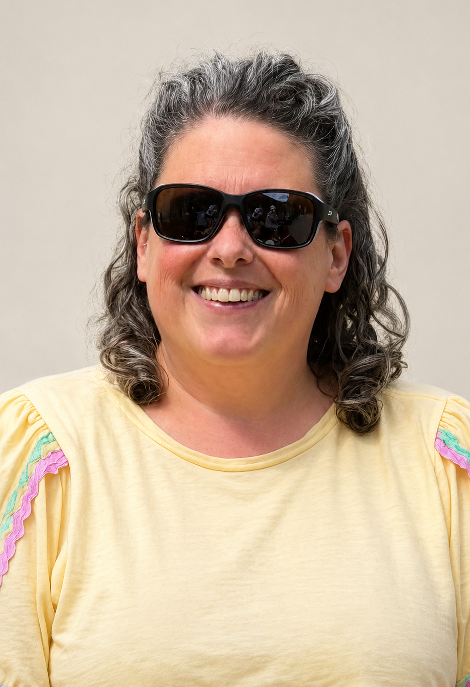 A head shot of Beth, a Tyne and Wear SLC member. She is stood against a cream background and has dark, shoulder-length, wavy hair. She is wearing dark sunglasses and a pale yellow t-shirt. She is smiling at the camera.