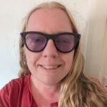 Close-up portrait of Nikki, South Yorkshire SLC member. Nikki has long, light coloured hair and is wearing dark framed sunglasses. The photo is taken indoors against a light coloured wall. Nikki is smiling at the camera.