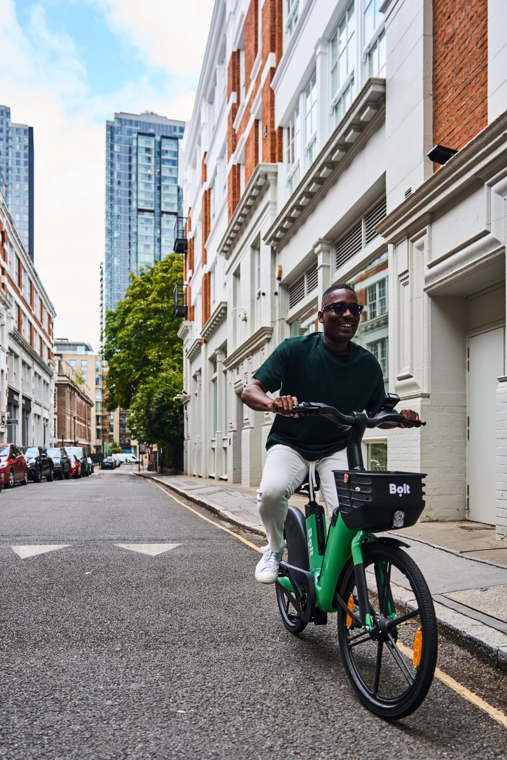 A male, wearing sunglasses, rides a green Bolt e-bike along a quiet city street lined with white and red-brick buildings. The bike has a black front basket displaying the Bolt logo. Modern high-rise buildings and trees can be seen in the background.