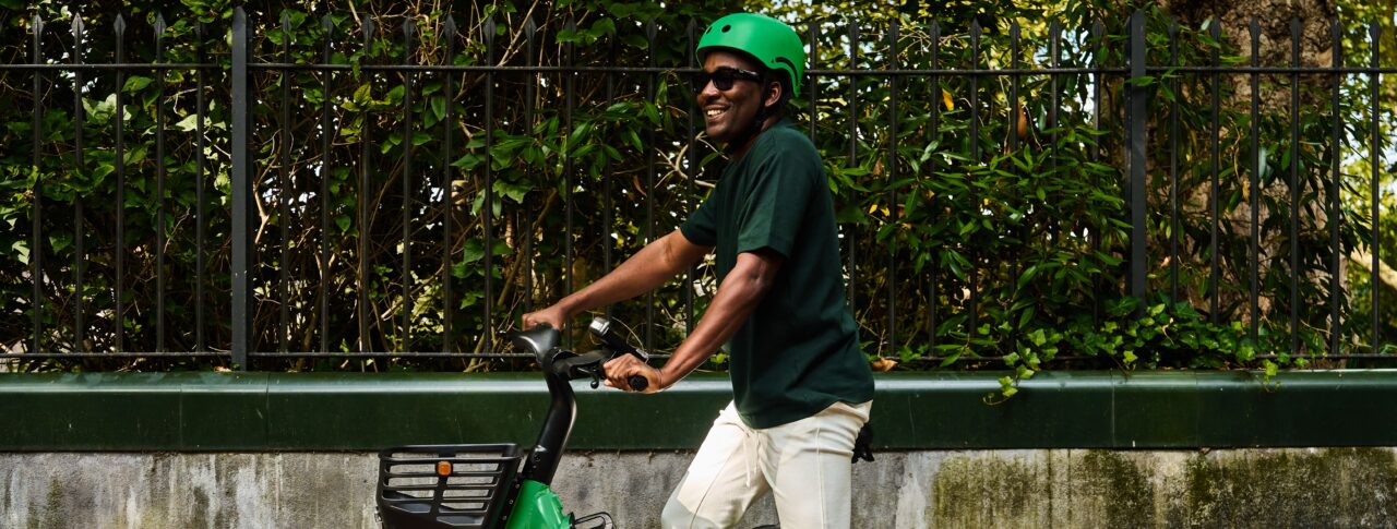 A person wearing a bright green cycle helmet and dark sunglasses sits on a green Bolt e-bike with a front basket, beside a low wall and black metal fence. Green foliage fills the background, and scattered leaves are visible on the ground. The rider is wearing a bright green helmet, a dark green T-shirt, light-coloured trousers and white trainers.