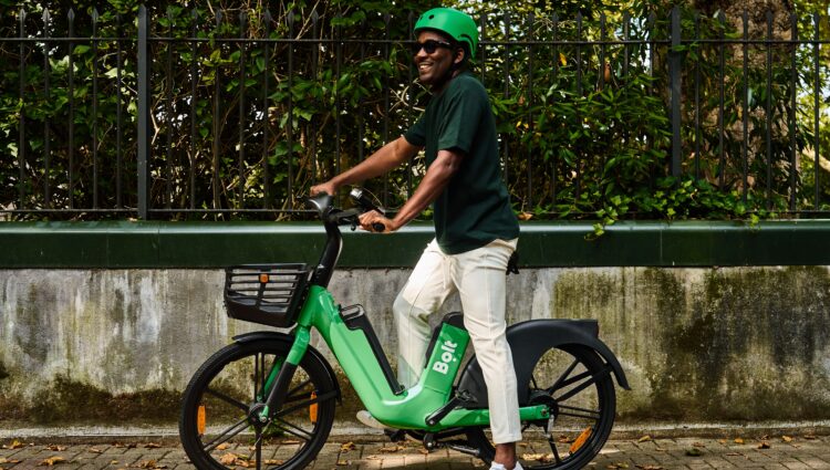 A person wearing a bright green cycle helmet and dark sunglasses sits on a green Bolt e-bike with a front basket, beside a low wall and black metal fence. Green foliage fills the background, and scattered leaves are visible on the ground. The rider is wearing a bright green helmet, a dark green T-shirt, light-coloured trousers and white trainers.