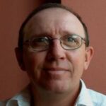 Headshot of Geoff Smith, Sussex SLC member. Geoff wears thin metal-framed glasses and a white collared shirt. Geoff faces the camera against pinky-brown walls.