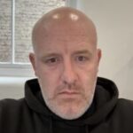 Headshot of Josh Brown, Sussex SLC member. Josh has a shaved head and a short, grey beard. He is wearing a black hoodie and looking at the camera.