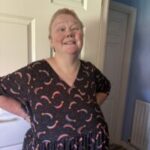 Photo of Justine Fallis, Surrey SLC member. Justine stands indoors infront of a white pannelled door. She is wearing a navy blue, patterned dress and is smiling at the camera.