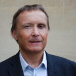 Head-and-shoulders portrait of a Surrey SLC member, Steve, wearing a navy blazer and light blue collared shirt, standing in front of a light stone wall. Steve is facing the camera against a plain background.