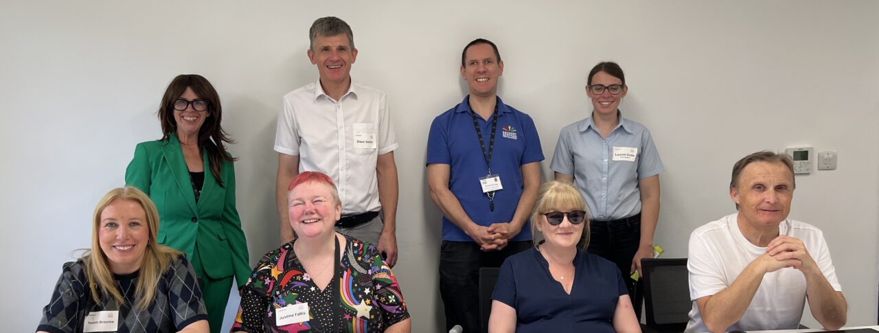 Surrey Sight Loss Council volunteers and representatives from partner organisations Sensory Services by Sight for Surrey and Surrey Coalition gather around a meeting table during the launch of Surrey Sight Loss Council. Refreshments and two celebration cakes are visible on the table. Everyone is facing the camera, smiling.