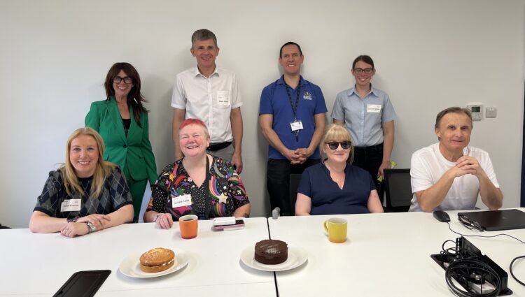 Surrey Sight Loss Council volunteers and representatives from partner organisations Sensory Services by Sight for Surrey and Surrey Coalition gather around a meeting table during the launch of Surrey Sight Loss Council. Refreshments and two celebration cakes are visible on the table. Everyone is facing the camera, smiling.