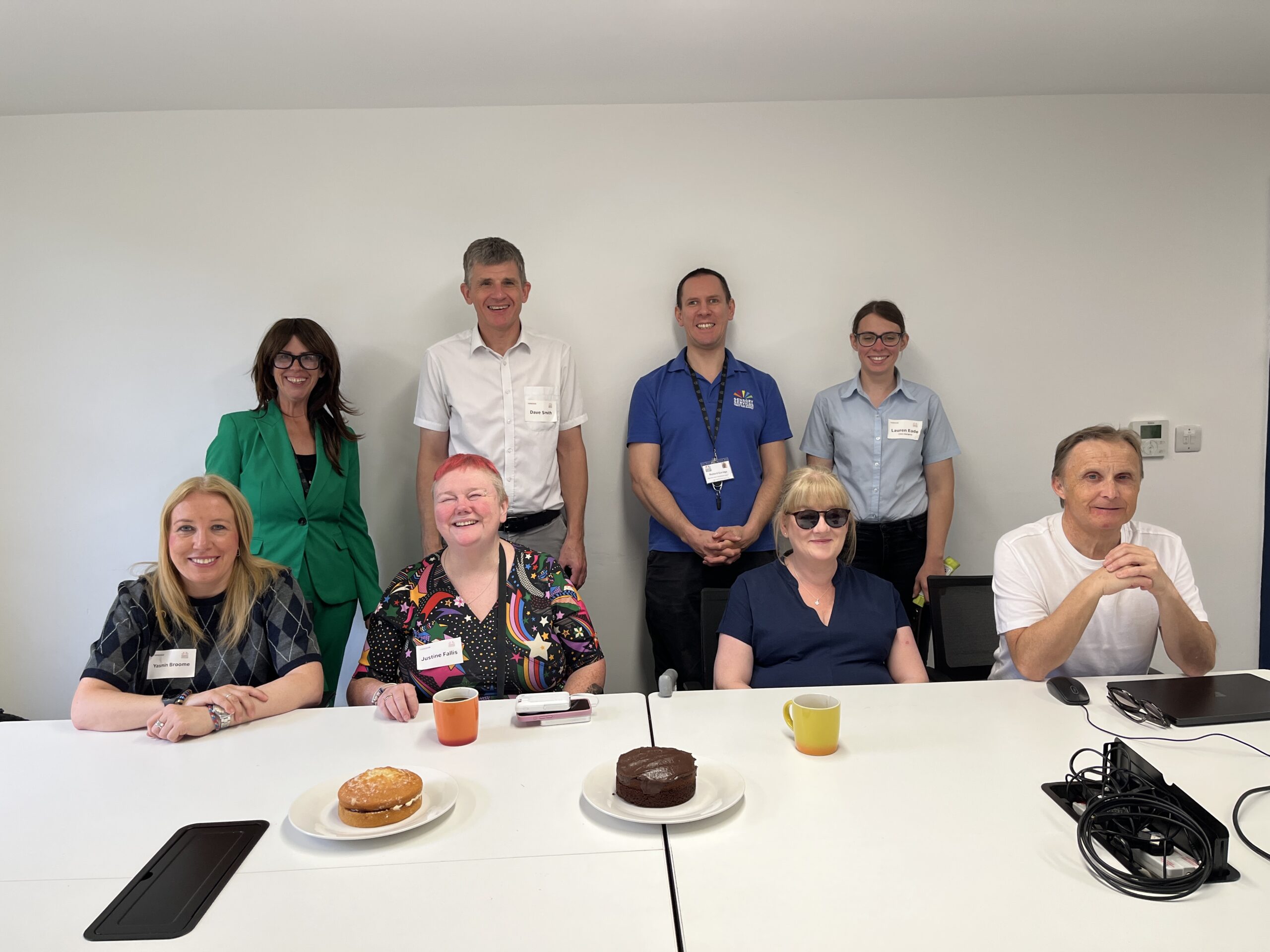 Surrey Sight Loss Council volunteers and representatives from partner organisations Sensory Services by Sight for Surrey and Surrey Coalition gather around a meeting table during the launch of Surrey Sight Loss Council. Refreshments and two celebration cakes are visible on the table. Everyone is facing the camera, smiling.