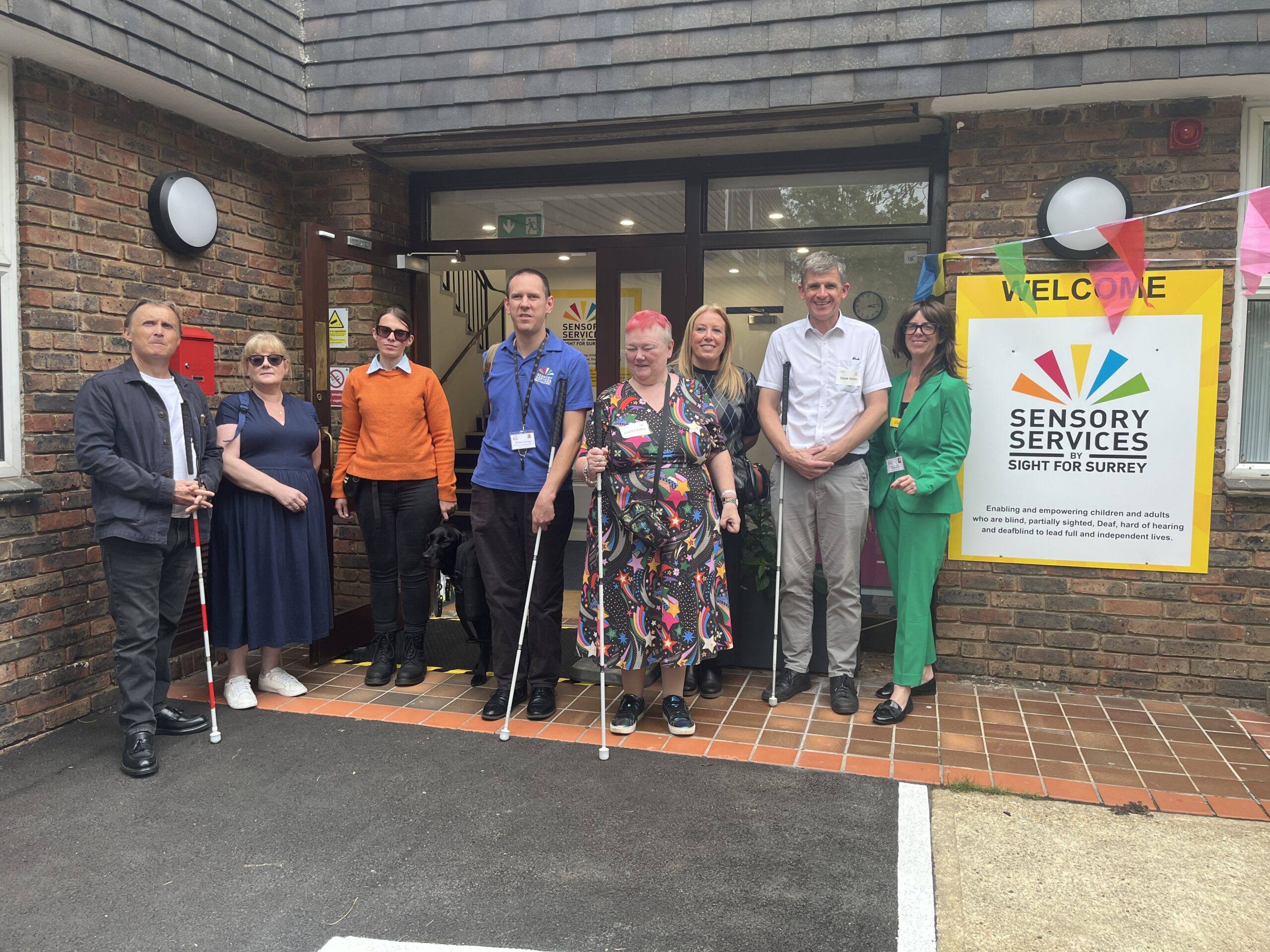 Surrey Sight Loss Council volunteers stand outside Sensory Services by Sight for Surrey with representatives from Sensory Services by Sight for Surrey and Surrey Coalition following the launch of Surrey Sight Loss Council. Several attendees are holding long canes.