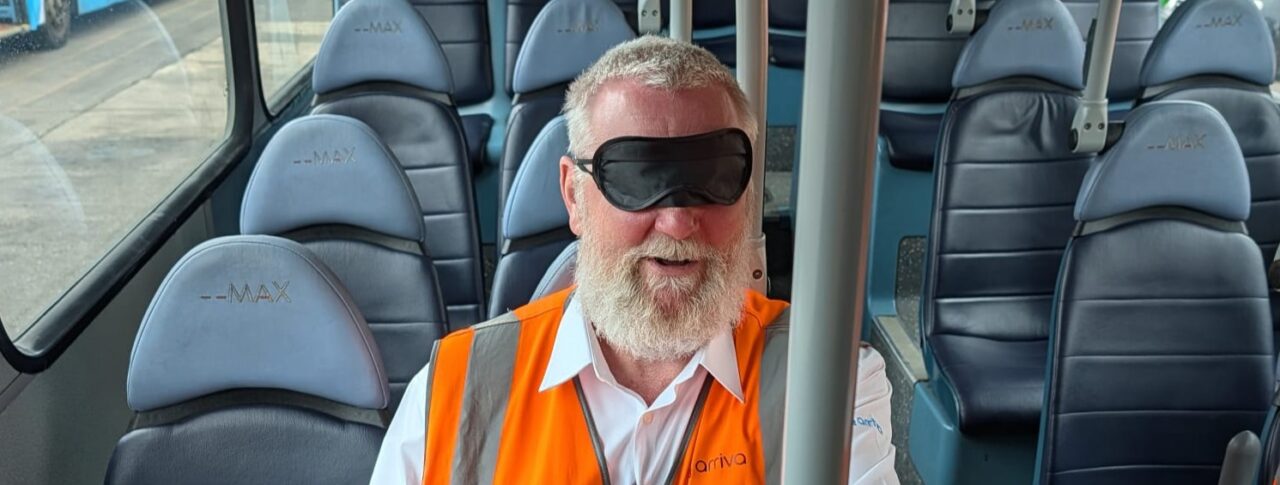 A male bus driver, wearing a high-visibility orange vest and a blindfold, sits on a bus during a vision awareness training exercise. He is seated among rows of blue passenger seats and holds a vertical grab pole to help with orientation.