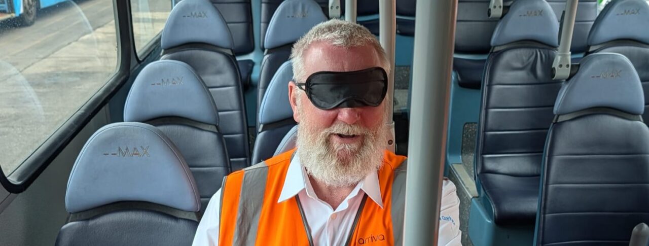 A male bus driver, wearing a high-visibility orange vest and a blindfold, sits on a bus during a vision awareness training exercise. He is seated among rows of blue passenger seats and holds a vertical grab pole to help with orientation.
