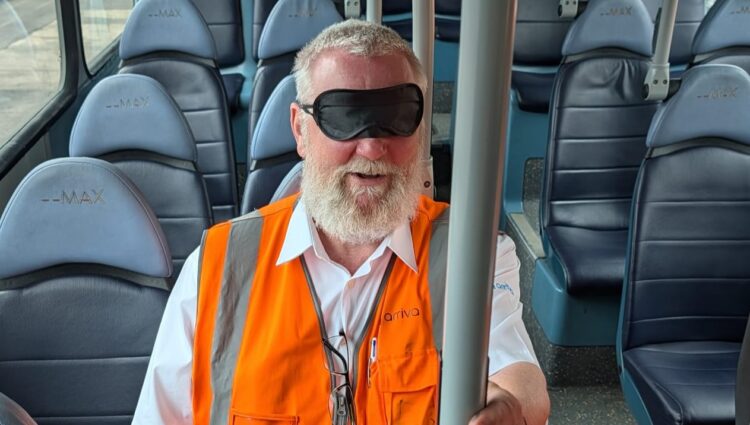 A male bus driver, wearing a high-visibility orange vest and a blindfold, sits on a bus during a vision awareness training exercise. He is seated among rows of blue passenger seats and holds a vertical grab pole to help with orientation.