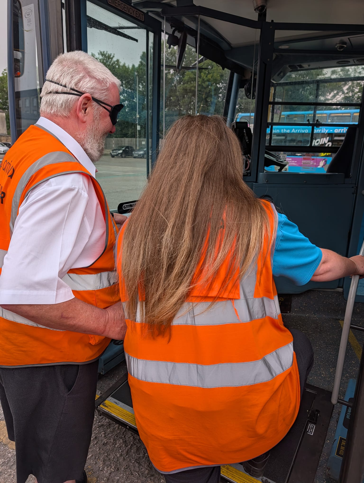 A practical bus driver awareness training exercise taking place at the entrance of a stationary bus. Two participants wearing high-visibility orange vests practise sighted guiding techniques, with one participant wearing a blindfold while boarding the vehicle.