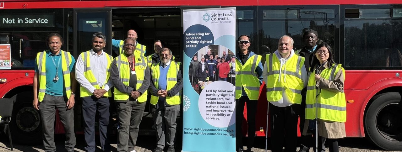 Sight Loss Council volunteers, Arriva bus drivers and managers, TfL representatives, and staff members stand together in front of a red double-decker bus at Arriva's Clapton Bus Garage. The group includes George Marcar, TfL Driver Communications Manager, and Brian Gordon, TfL Travel Mentor Manager. A Sight Loss Councils banner is displayed in the centre of the group, and several participants are wearing high-visibility vests