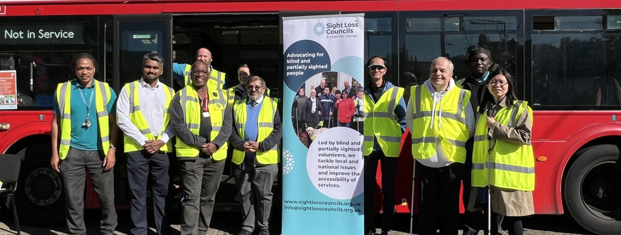 Sight Loss Council volunteers, Arriva bus drivers and managers, TfL representatives, and staff members stand together in front of a red double-decker bus at Arriva's Clapton Bus Garage. The group includes George Marcar, TfL Driver Communications Manager, and Brian Gordon, TfL Travel Mentor Manager. A Sight Loss Councils banner is displayed in the centre of the group, and several participants are wearing high-visibility vests