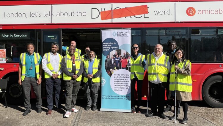 Sight Loss Council volunteers, Arriva bus drivers and managers, TfL representatives, and staff members stand together in front of a red double-decker bus at Arriva's Clapton Bus Garage. The group includes George Marcar, TfL Driver Communications Manager, and Brian Gordon, TfL Travel Mentor Manager. A Sight Loss Councils banner is displayed in the centre of the group, and several participants are wearing high-visibility vests