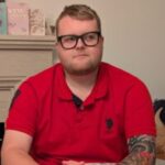 Photo of Harvey, Lancashire SLC member. Harvey is seated at a table with hands clasped. He is wearing black-framed glasses and a red polo shirt, with a patterned tattoo visible on one forearm. Greeting cards and a fireplace are visible in the background.