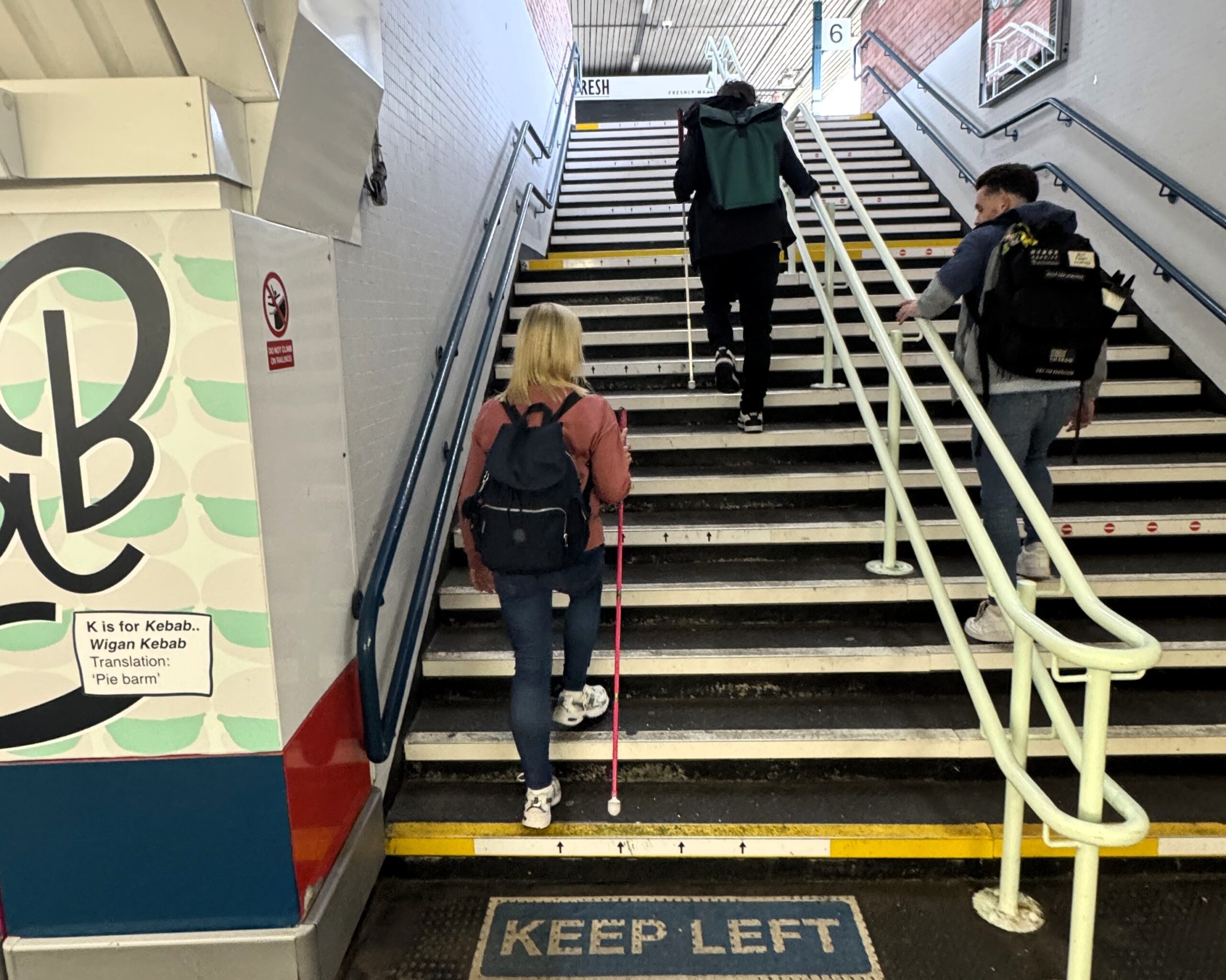 Kelly Barton, Engagement Manager for the North West, a SLC volunteer, and David Parfett, SLC Coordinator for North West England, walk up a staircase in a railway station. Handrails run along both sides and the centre of the staircase, with high-contrast step edges and a "KEEP LEFT" marking visible at the bottom.