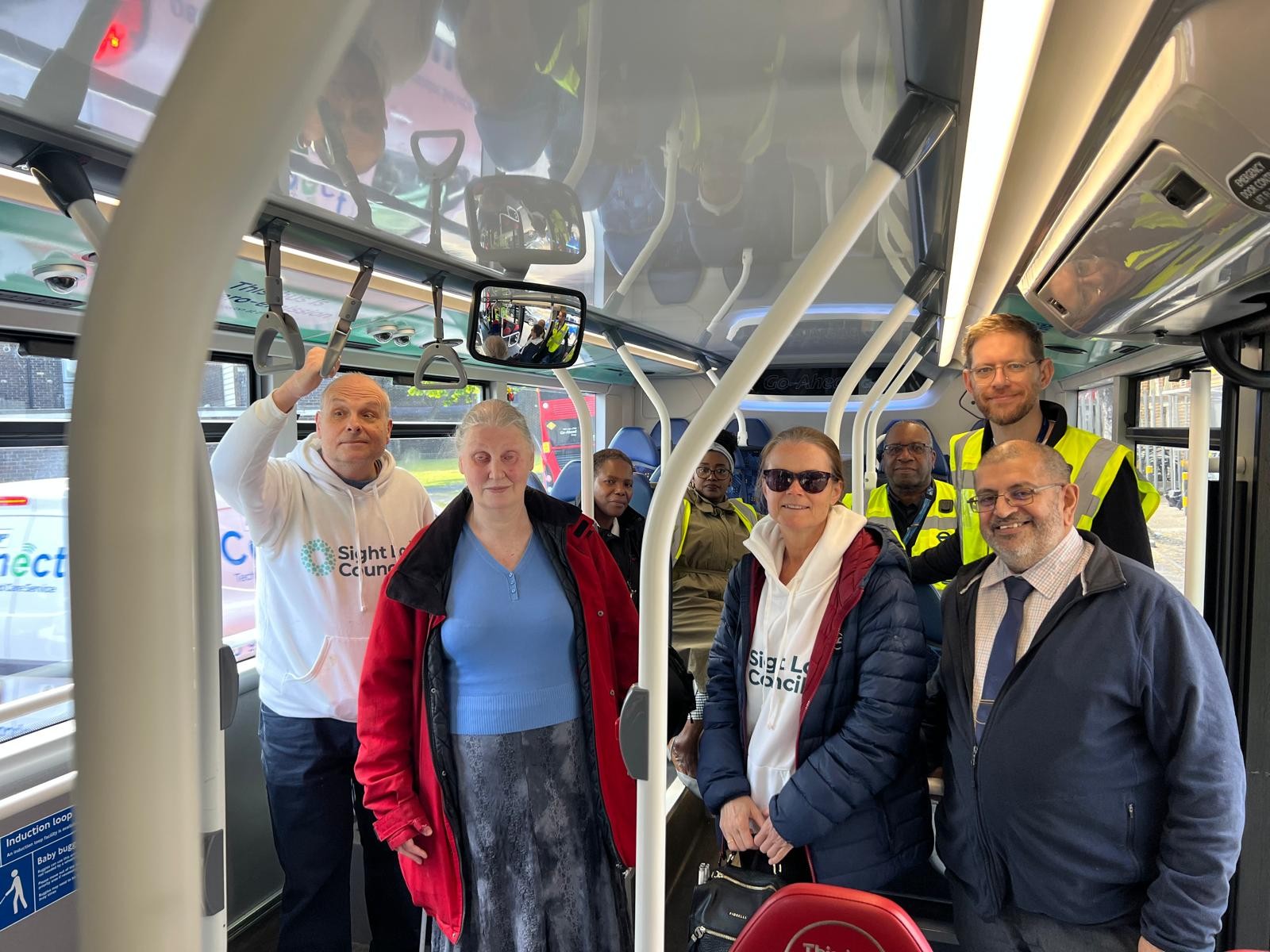 Liam O'Carroll, Interim Engagement Manager for London Sight Loss Councils, stands on board a bus with South West London Sight Loss Council volunteers Mary and Vicky. George Marcar, Driver Communications Manager at TfL, Brian Gordon, Travel Mentor Manager, and staff from Sutton Bus Garage are seated behind them during a bus driver vision awareness session.