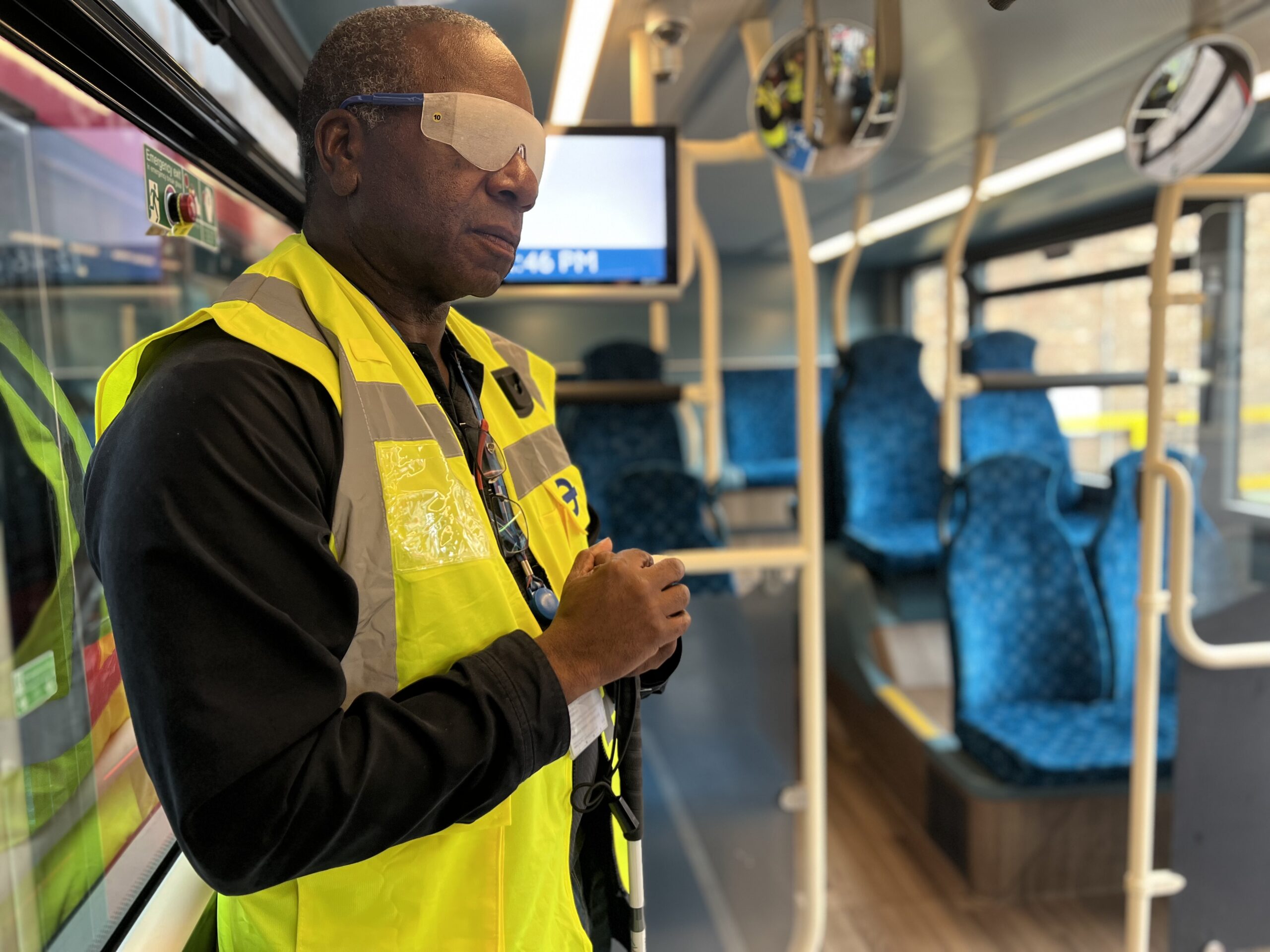 Brian Gordon, TfL Travel Mentor Manager, stands inside a stationary bus wearing a yellow high-visibility jacket and light perception-only simulation glasses as part of a sight loss awareness exercise. Blue passenger seats, handrails and bus mirrors are visible in the background.
